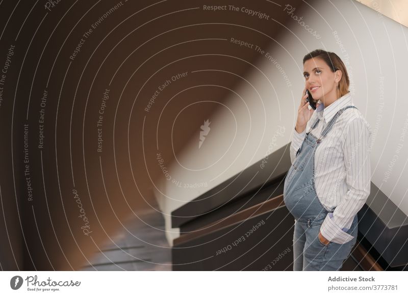 Lächelnde schwangere Frau, die in einem Gebäude mit einem Mobiltelefon spricht sprechen Smartphone Schwangerschaft Bauch modern Treppenhaus benutzend Reling