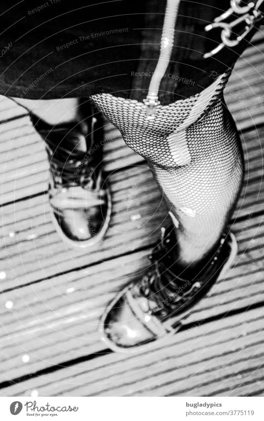 Frau von der Hüfte abwärts im Stehen fotografiert. Sie trägt einen schwarzen Rock mit einem auffälligen Reißverschluss und Ketten, eine Netzstrumpfhose mit Laufmaschen und Löchern und derbe schwarze Schuhe. Der Fokus liegt auf der großen Laufmasche.