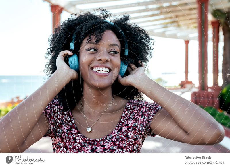 Lächelnde schwarze Frau genießt Musik auf der Promenade Drahtlos Kopfhörer zuhören genießen Freude sorgenfrei Sommer Spazierweg ethnisch Afroamerikaner