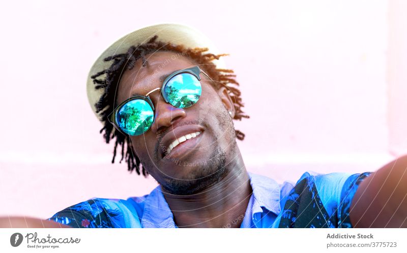 Lächelnder schwarzer Mann nimmt Selfie in der Stadt Smartphone tropisch Hemd drucken Stil trendy Freude benutzend männlich ethnisch Afroamerikaner Gebäude