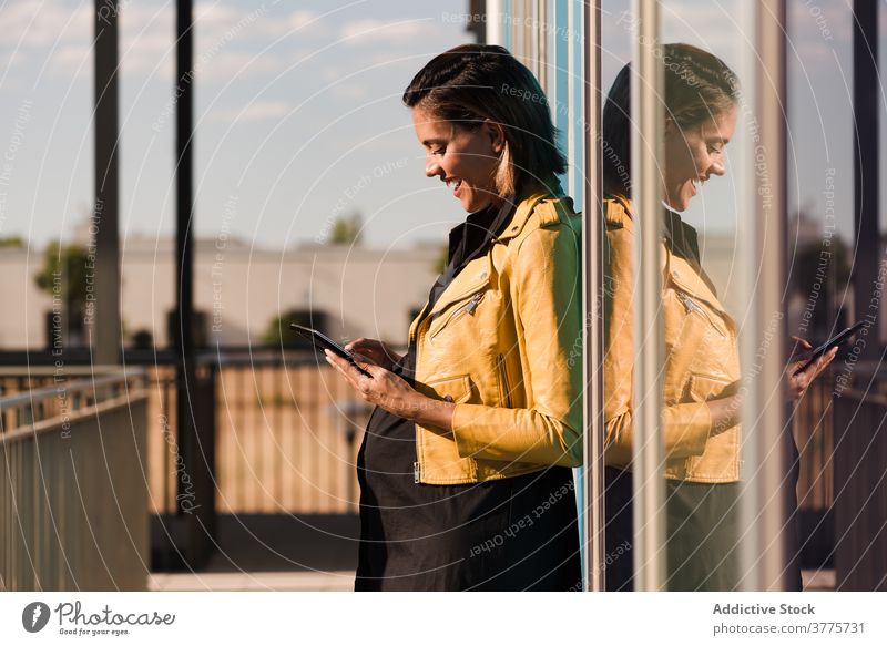 Lächelnde schwangere Frau liest Nachrichten auf dem Smartphone Talkrunde soziale Netzwerke Großstadt Glaswand Gebäude Sonnenlicht benutzend Bauch heiter Wand