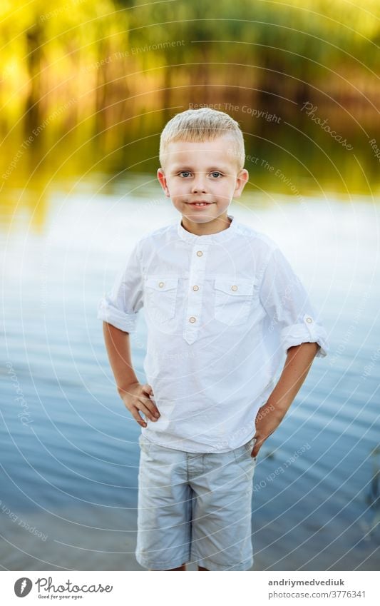 Porträt eines glücklichen und schönen kleinen Jungen mit blonden Haaren und weißem Hemd, Glückliche Kindheit. Positive Emotion. Porträt in der Nähe des Flusses in der Natur