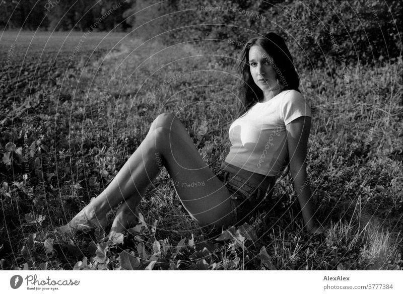 Schwarzweiß- Portrait einer jungen Frau, die vor einem Wald am Feldrand im Gras sitzt schön nah fit anmutig Haut Gesicht blond schauen direkt langhaarig schlank