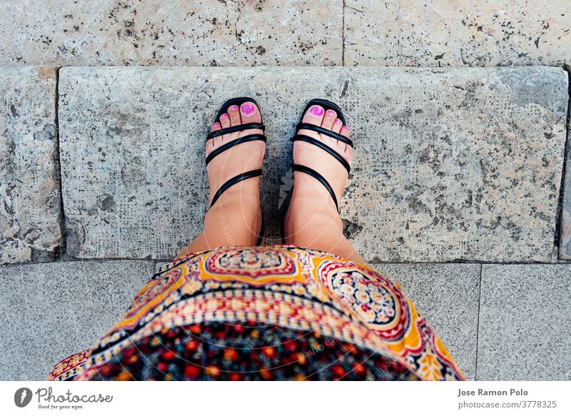 Frauenfüße in Ich-Perspektive mit fuchsiafarben lackierten Nägeln und schwarzen Schuhen, in marokkanischer Stammeskleidung, auf grauen Steinfliesen. ethnisches Konzept