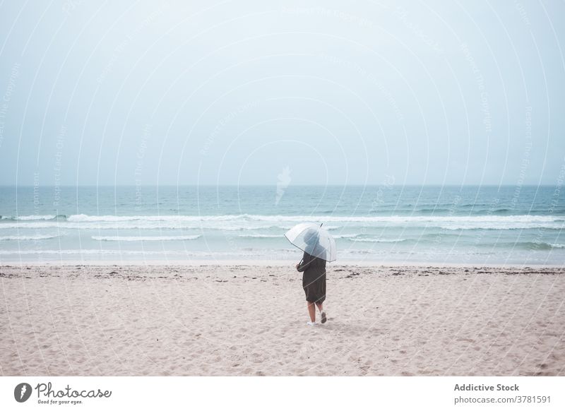 Anonyme Person mit Regenschirm, die am Sandstrand spazieren geht Strand MEER allein reisen Meer einsam stürmisch bedeckt Reisender Ufer Küste Natur winken