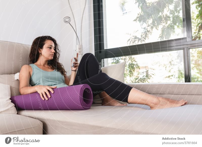 Ruhige Frau entspannt auf Sofa mit Smartphone und Yogamatte Browsen heimwärts Unterlage Lügen benutzend Wochenende sich[Akk] entspannen Sportbekleidung