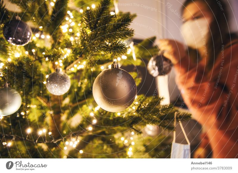Junge Frau schmückt einen Weihnachtsbaum mit medizinischer Sicherheitsmaske für Covid-19, Coronavirus und Weihnachtskonzept, schöner Feiertag Neujahr heimwärts
