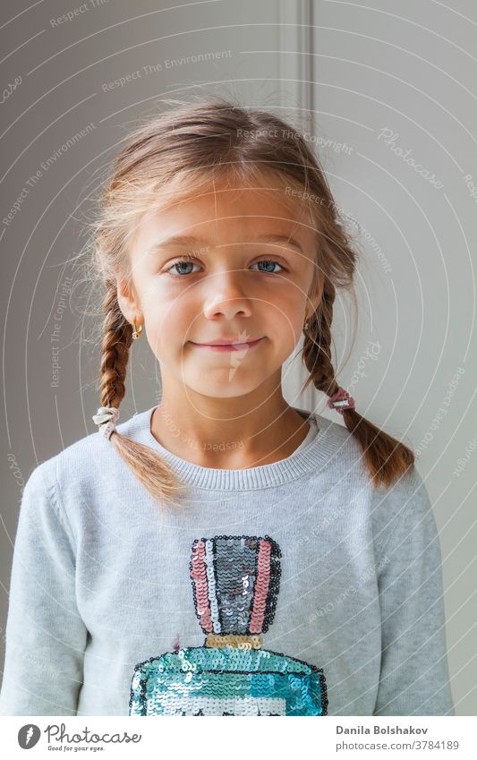 Nahaufnahme eines schönen сaucasian kleinen Mädchens mit Zöpfen in einem gemütlichen Pullover, das mit einem charmanten Lächeln in die Kamera blickt und sich vor der hellen Fläche der Betonmauer postiert. Konzept der glücklichen Kindheit