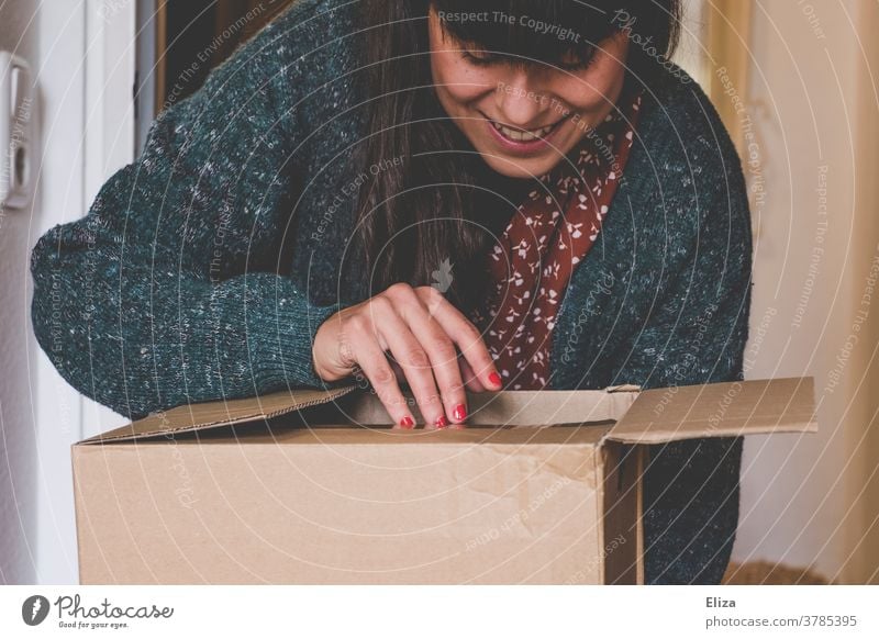 Eine junge Frau öffnet neugierig und voller Vorfreude ein Paket Onlineshopping auspacken Freude online bestellen Post Geschenk Überraschung lächeln Verpackung