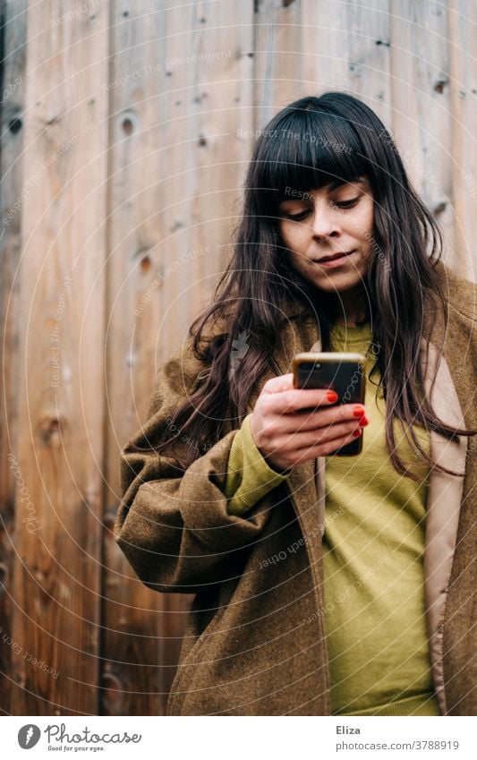 Junge Frau mit langen braunen Haaren und herbstlicher Kleidung lehnt an einer Holzwand und benutzt ihr Smartphone draußen Handy benutzen lange Haare jung