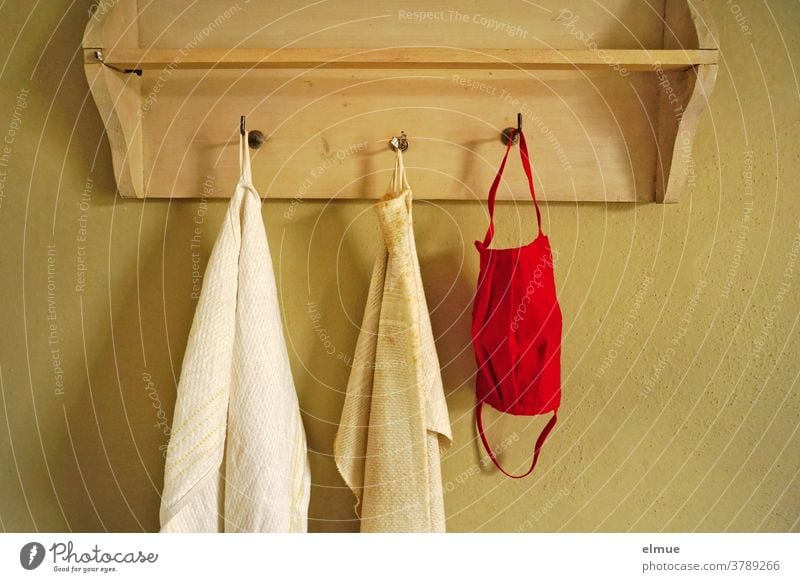 Eine rote Nasen- Mundschutzmaske hängt neben zwei alten, schmutzigen Handtüchern an einem der drei Haken einer Wandgarderobe aus Holz Handtuch Prävention Maske