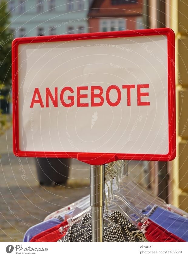 "Angebote" steht in großen roten Druckbuchstaben auf einem weißen Schild an einem Metall - Kleiderständer mit Bekleidung auf Kleiderbügeln inmitten der Kleinstadt