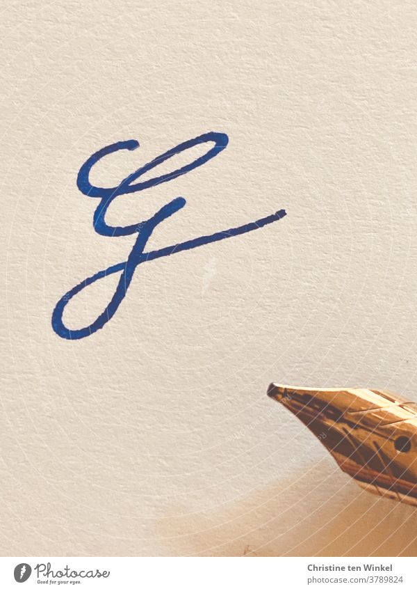 Der verschnörkelte Buchstabe G  auf hellem strukturiertem Papier, geschrieben mit blauer Tinte. Am rechten Bildrand sieht  man die goldfarbene Spitze des Füllfederhalters