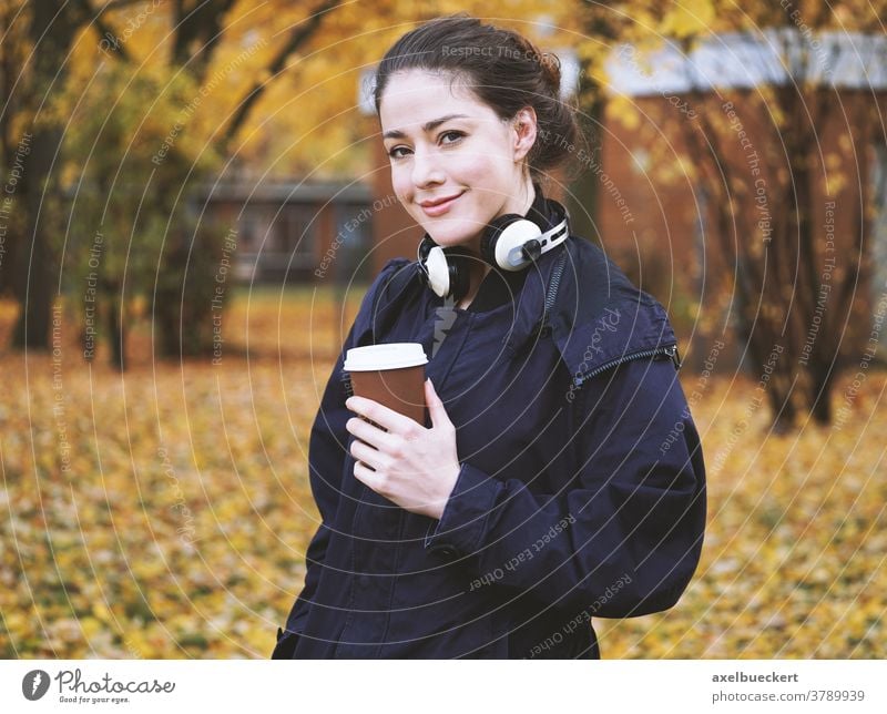 junge Frau mit Kaffee zum Mitnehmen und Kopfhörern im Herbst offen im Freien außerhalb echte Menschen fallen heiß trinken kalt Wetter Straße Spaziergang Park