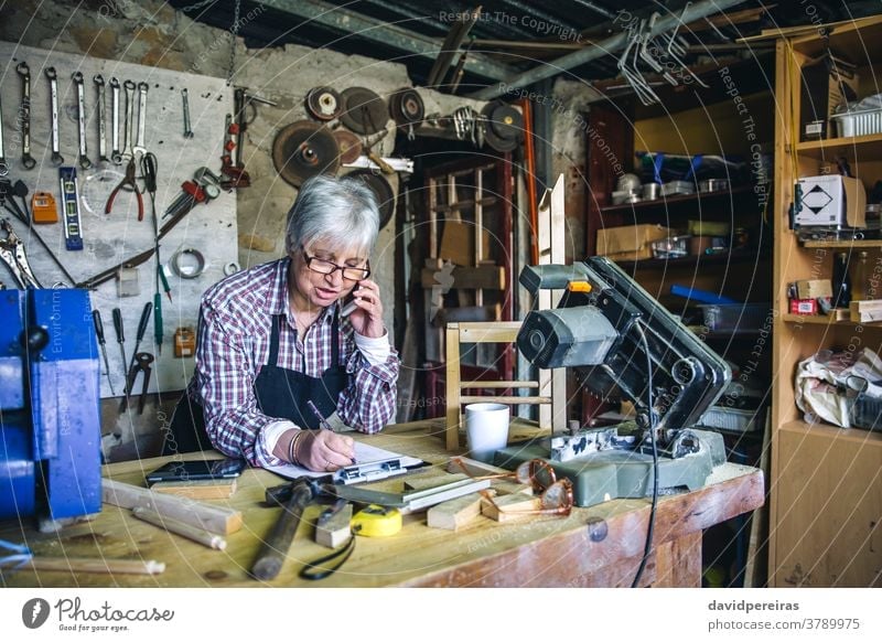 Zimmermannsfrau in ihrer Werkstatt Schreinerei Frau Zimmerer Senior sprechend Mobile Handy maßgefertigt Business reif schreibend Holz Menschen alt Kaukasier
