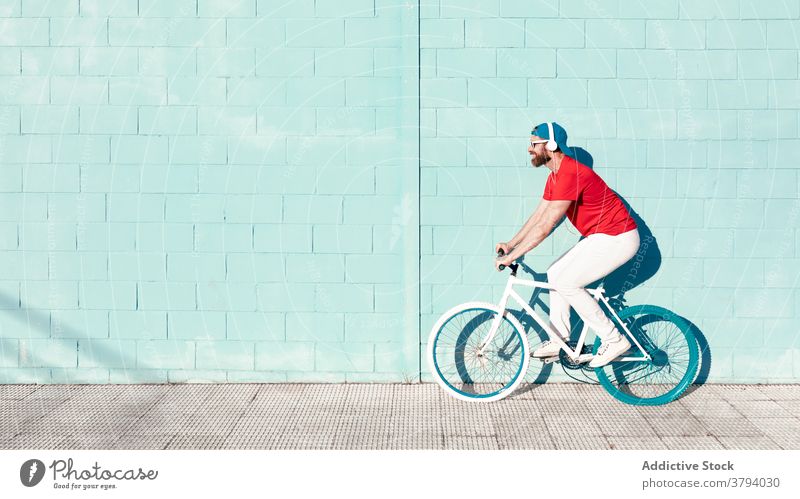 Junger Mann fährt Fahrrad und hört Musik in Kopfhörern auf der Stadtstraße Mitfahrgelegenheit Straße zuhören Radfahrer aktiv Trick trendy Großstadt Gebäude