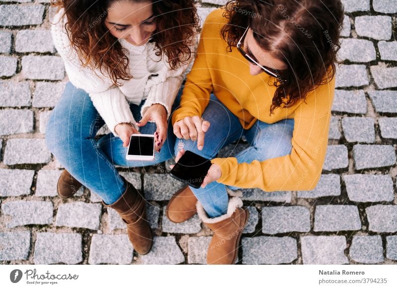 Selektiver Schwerpunkt. Zwei schöne Frauen sitzen auf der Straße und benutzen ihre Telefone. Beide sehen sich etwas auf dem Handy an. Technischer Lebensstil