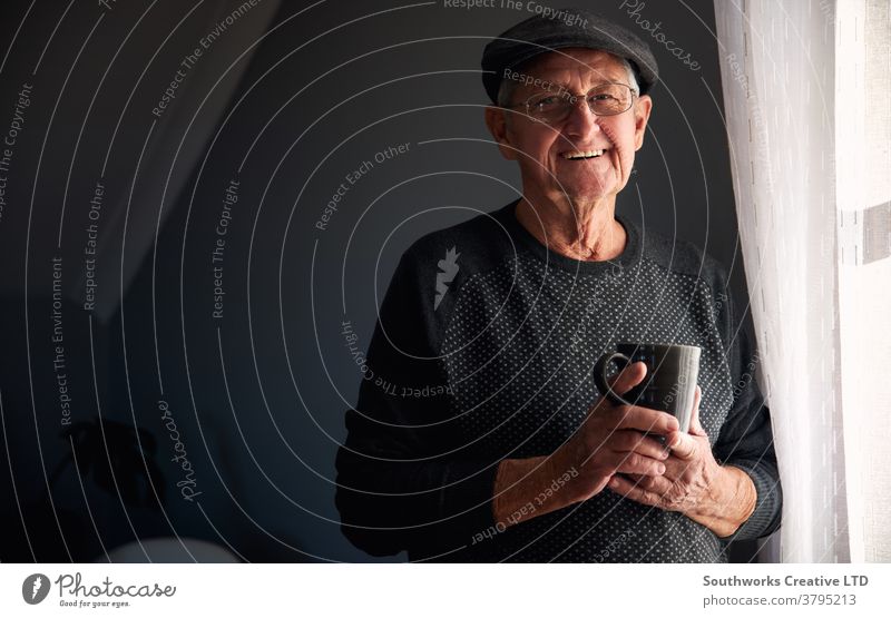 Porträt eines älteren Mannes mit Mütze, der entspannt am Fenster steht und zu Hause ein heisses Getränk zu sich nimmt Senior Senioren in den Ruhestand getreten