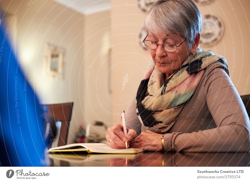 Ältere Frau zu Hause sitzt am Tisch und schreibt in Notizbuch oder Journal Senior Senioren schreibend Notebook Tagebuch Liste erstellen Notizen machen