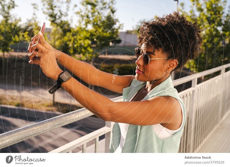 Stilvolle ethnische Frau in Sonnenbrille unter Selfie in der Nähe von Zaun trendy Glück Sommer Afro-Look Lächeln heiter jung Afroamerikaner schwarz positiv