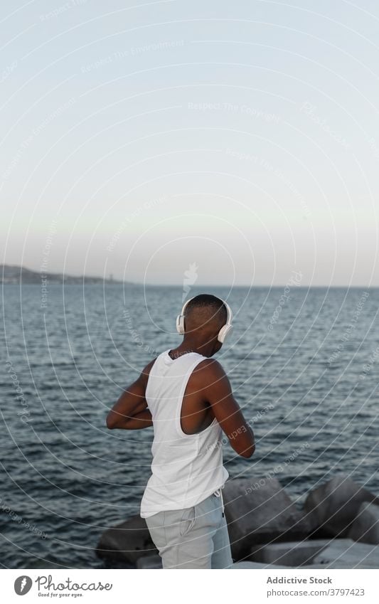 Schlanker schwarzer Mann, der sich beim Training an der Strandpromenade dehnt Läufer Hafengebiet Aufwärmen Kopfhörer Dehnung Energie Konzentration Wohlbefinden