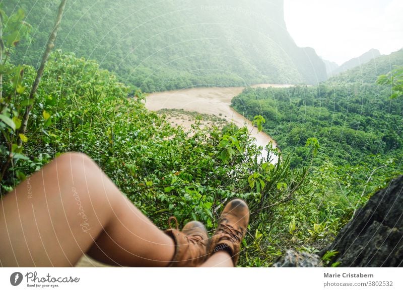 Die Beine eines Wanderers ruhen auf einem Felsvorsprung mit Blick auf den Fluss und den üppigen Wald im Dorf Nong Khiaw in Luang Prabang, Laos Nong Kiaw wandern