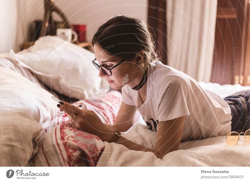 Hübsches Mädchen mit Brille liegt im Bett und schaut etwas auf dem Handy Frau jung Mobile Tippen schreibend Lügen Telefon heimwärts Funktelefon Lifestyle