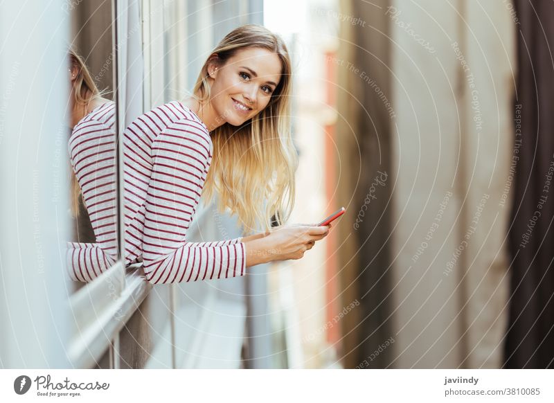 Junge Frau lehnt sich mit einem Smartphone aus dem Fenster ihres Hauses. Erwachsener attraktiv Hintergrund schön Schönheit schwarz Anruf lässig Kaukasier Zelle
