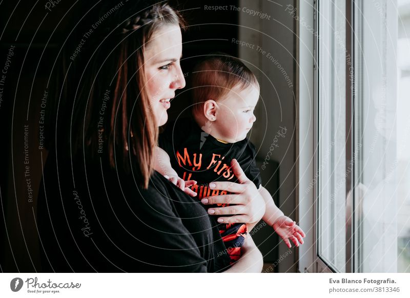 junge Mutter und kleines Mädchen zu Hause, die durch das Fenster schauen. Kleines Mädchen in Halloween-Kleidung. Familienleben im Haus Baby heimwärts Liebe