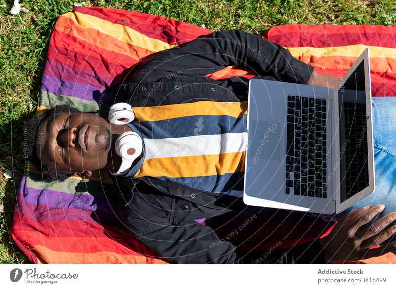 Sorgloser schwarzer Mann auf Decke liegend mit Laptop im Park müde freiberuflich schlafen Mittagsschlaf abgelegen Unternehmer männlich ethnisch Afroamerikaner