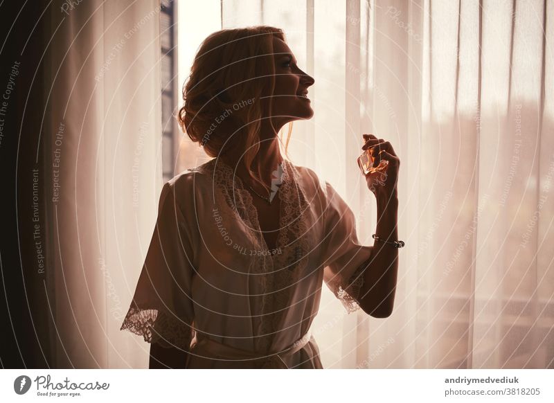 Ein Mädchen in einem weißen Satinmantel mit Ausschnitt und Manikürfranzösisch hält einen Flakon mit Parfüm und Spritzern am Handgelenk. Morgen der Braut im Haus am Fenster