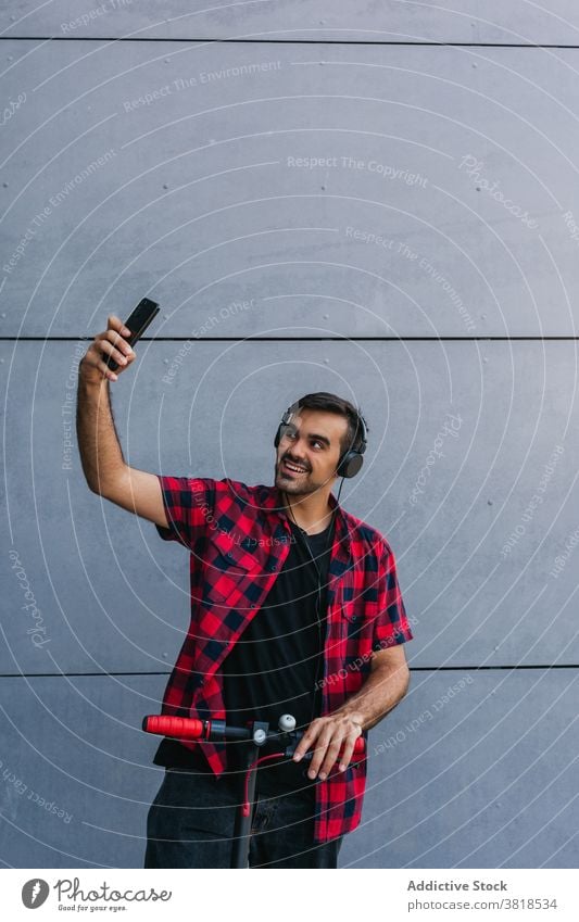Fröhlicher Mann auf Elektroroller nimmt Selfie in der Stadt Hipster Tretroller elektrisch Stil kariertes Hemd Kopfhörer Großstadt unterhalten männlich