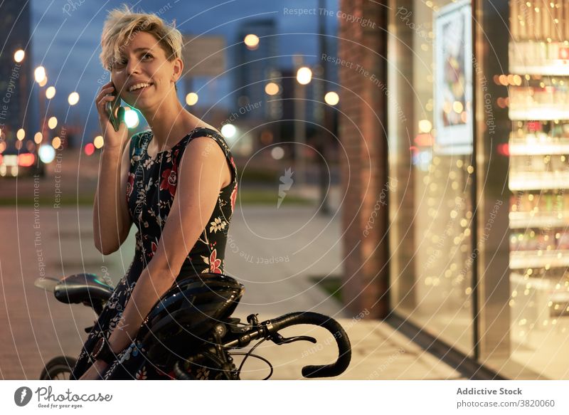 Junge Frau spricht am Telefon in der Nähe von Geschäft und Fahrrad Kleid Abend Spaziergang Mädchen jung Reiten schön Großstadt Nacht Lichter Gebäude Glas