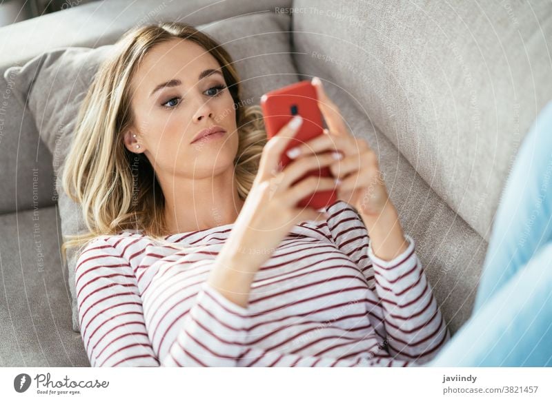 Junge blonde Frau benutzt ihr Smartphone auf dem Sofa liegend. heimwärts Mädchen Mobile Telefon jung weiß Funktelefon Menschen im Innenbereich Liege eine