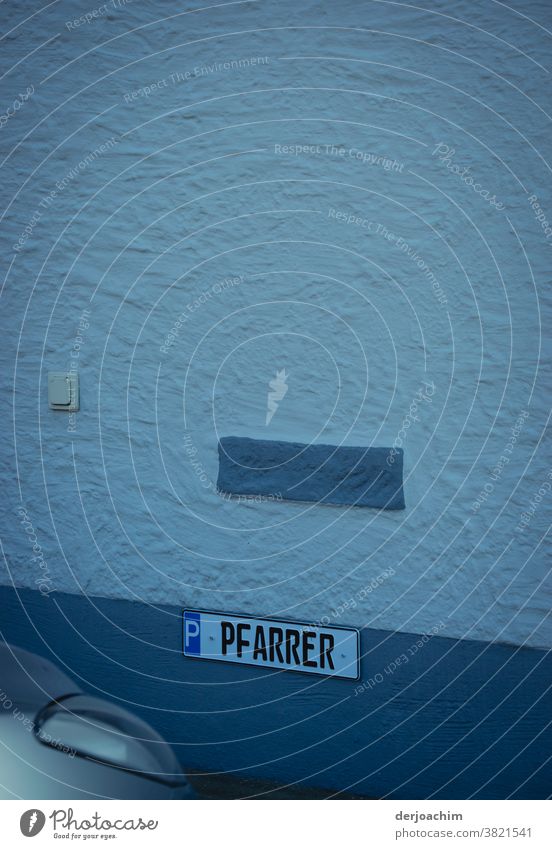 Parkplatz für den PFARRER. Der ist leider von einem unbekannten PkW besetzt. Das Schild ist an einer weißen mit blau abgestuften Wand befestigt . Im unteren Bildrand links, ist ein Scheinwerfer zu erkennen.