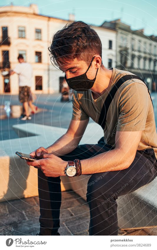 Junger Mann sitzt im Stadtzentrum, benutzt ein Smartphone und trägt die Gesichtsmaske, um eine Virusinfektion zu vermeiden Kaukasier Gespräch covid-19 Lifestyle