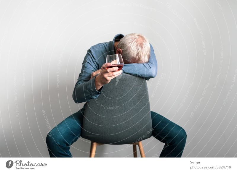 Depressiver Mann sitzt auf einem Sessel mit dem Kopf auf einem Arm und einem Glas Cognac in der Hand Depression Verzweiflung Einsamkeit traurig unglücklich