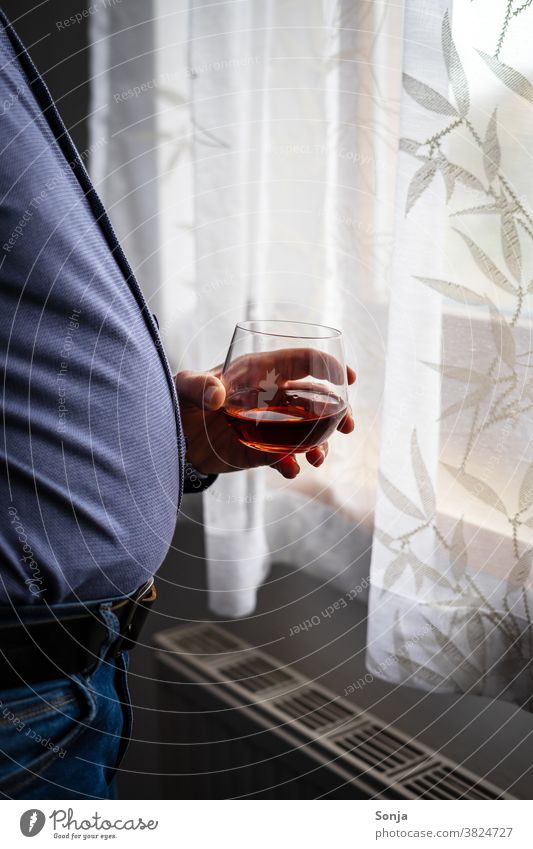 Übergewichtiger Mann mit einem Glas Cognac steht an einem Fenster Trinkglas Hand halten beobachten Einsamkeit zuhause zuhause bleiben dick Coronavirus