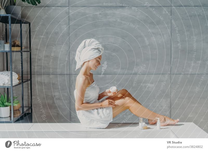 Horizontale Aufnahme einer entspannten jungen Europäerin trägt feuchtigkeitsspendende Körpercreme auf die Beine auf, in ein Badetuch gewickelt, hat ein zartes Lächeln, gesunde, erfrischte Haut nach dem Bad, posiert in einem gemütlichen Badezimmer.