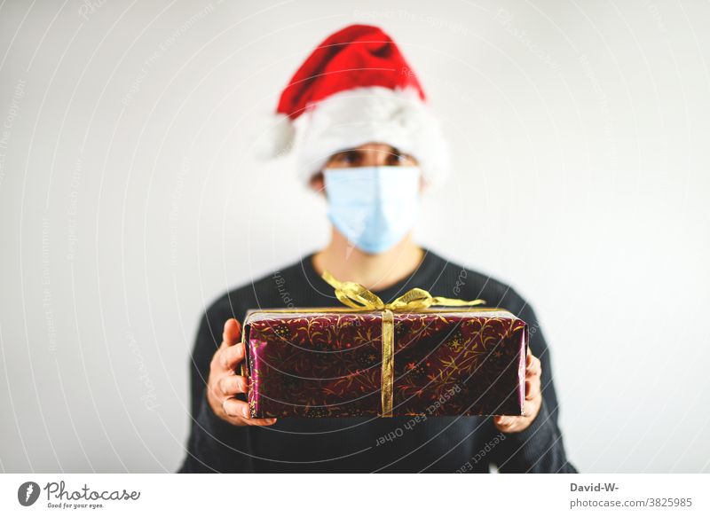 frohe Weihnachten - Mann hält unter Corona-Bedingungen Geschenk in den Händen Atemschutzmaske Mundschutz Nikolausmütze Bescherung pandemie Schutz Virus Schützen