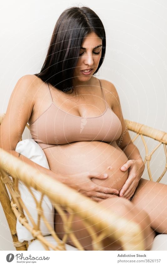Lächelnde schwangere Frau im Sessel zu Hause berühren Bauch Schwangerschaft gemütlich Dessous Unterwäsche sanft filigran Mutter Pflege Angebot sitzen erwarten