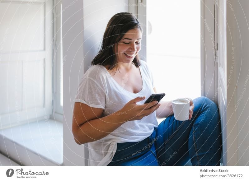 Mädchen sitzt am Fenster, trinkt Kaffee und benutzt ein Smartphone im Innenbereich heimwärts Haus Frau kaufen Küche Frühstück Morgen Kaukasier brünett Tisch