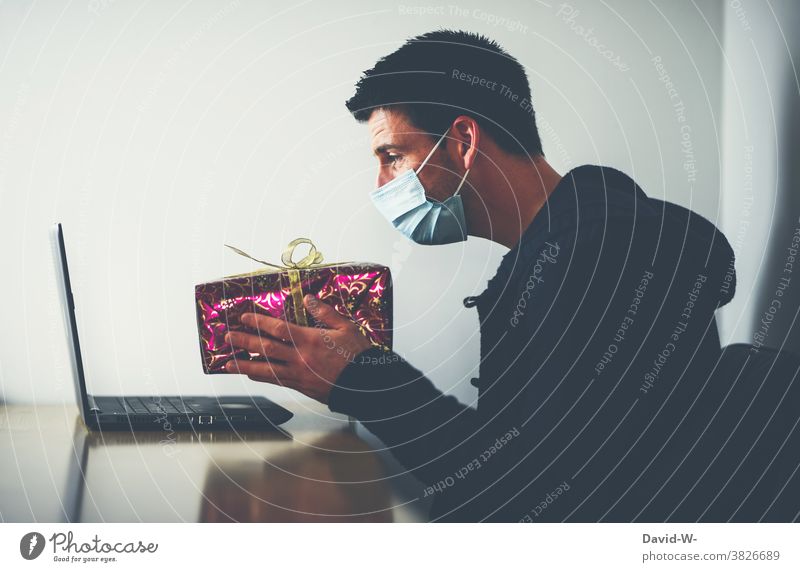 Corona und Weihnachten - Bescherung online - Mann sitzt mit Atemschutzmaske / Mundschutz am Laptop und hält ein Geschenk in die Webcam Quarantäne Pandemie