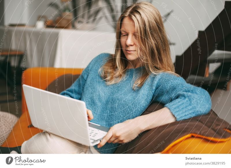 Junge Frau benutzt Laptop, checkt online auf dem Sofa sitzend E-Mail-Nachrichten. Hübsches Mädchen arbeitet am Computer, sieht sich ein Webinar an, lernt zu Hause