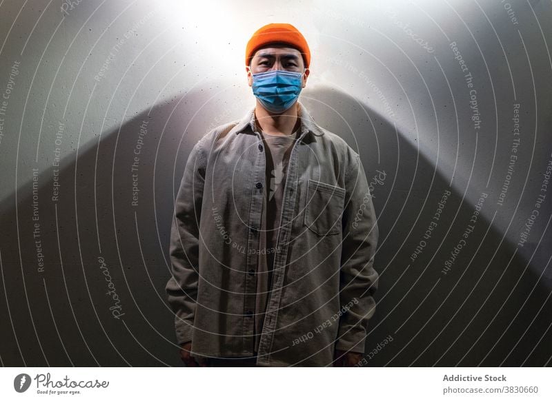 Ernster asiatischer Mann mit Gesichtsmaske steht in grauem Korridor Mundschutz Atemschutzgerät Konzentration nachdenklich besinnlich Windstille Deckel Mund