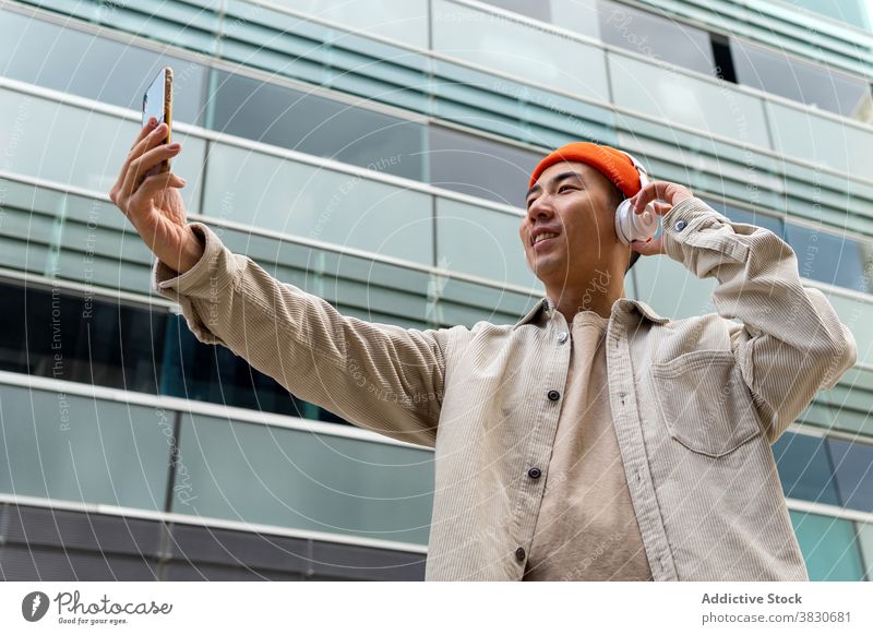 Fröhlich asiatischen Mann mit Videoanruf außerhalb modernen Gebäude Smartphone Gespräch reden Kopfhörer Talkrunde Freude Video-Chat Glück Kondominium Drahtlos