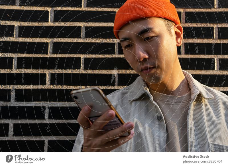 Verträumter asiatischer Mann, der sein Smartphone gegen eine Ziegelmauer nimmt Inhalt Gespräch Telefonanruf reden verträumt genießen Glück zuhören Gerät