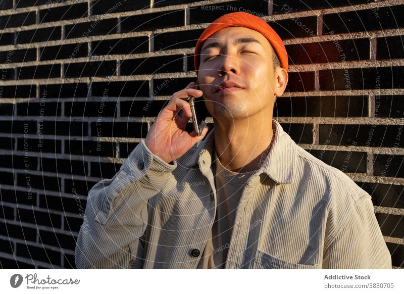 Verträumter asiatischer Mann, der sein Smartphone gegen eine Ziegelmauer nimmt Inhalt Gespräch Telefonanruf reden verträumt genießen Glück zuhören
