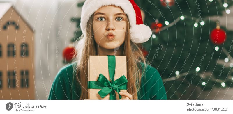 Ein schönes Mädchen mit überraschten Augen schaut in die Kamera mit einem Geschenk aus Kraftpapier, das mit einem grünen Band gebunden ist. Weihnachtsstimmung. Konzept für den Neujahrsfeiertag zu Hause.