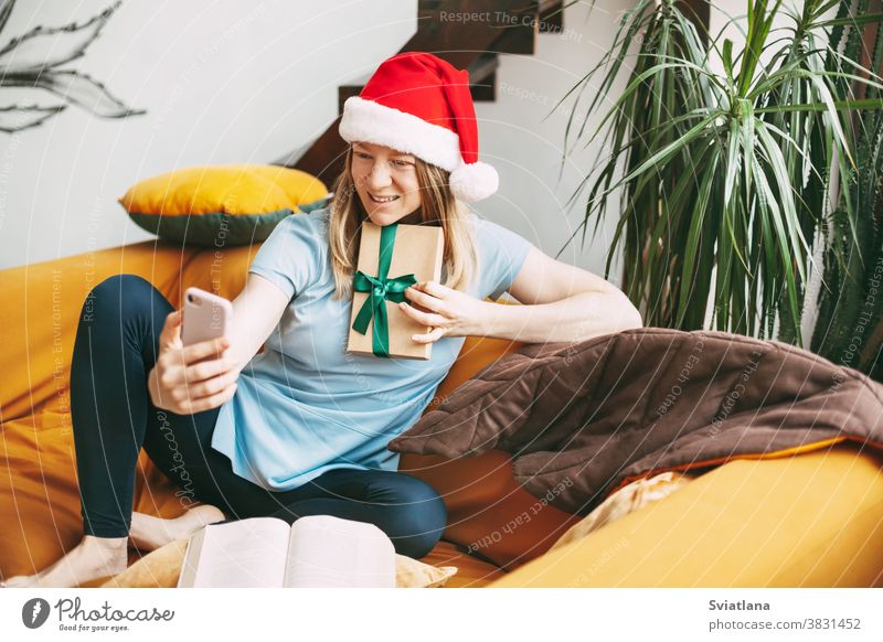 Ein lächelndes Mädchen mit Weihnachtshut sitzt auf dem Sofa und kommuniziert per Videolink mit Freunden und zeigt ihnen ihr Geschenk. Mädchen gratuliert Freunden und Familie zu den Feiertagen